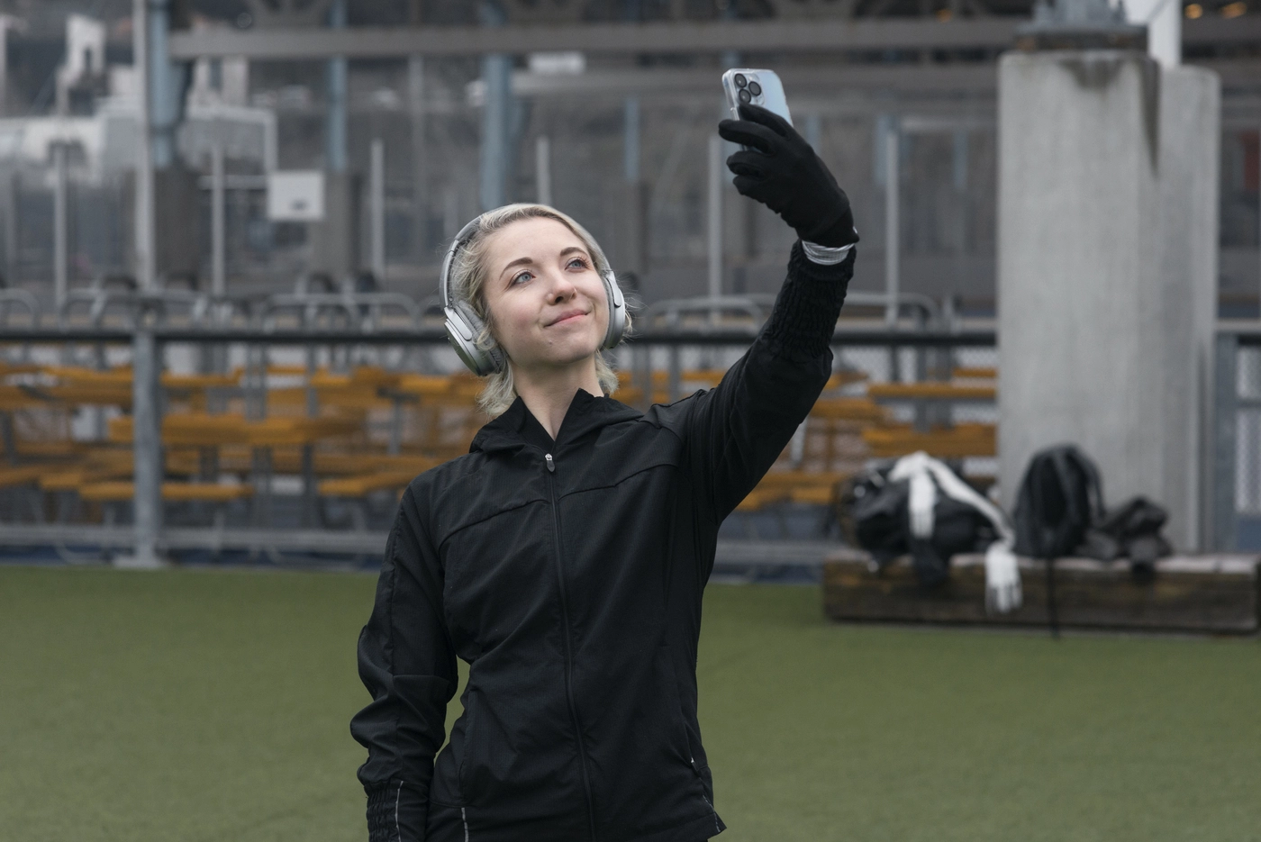 A person in a black jacket and headphones is taking a selfie with their phone outdoors